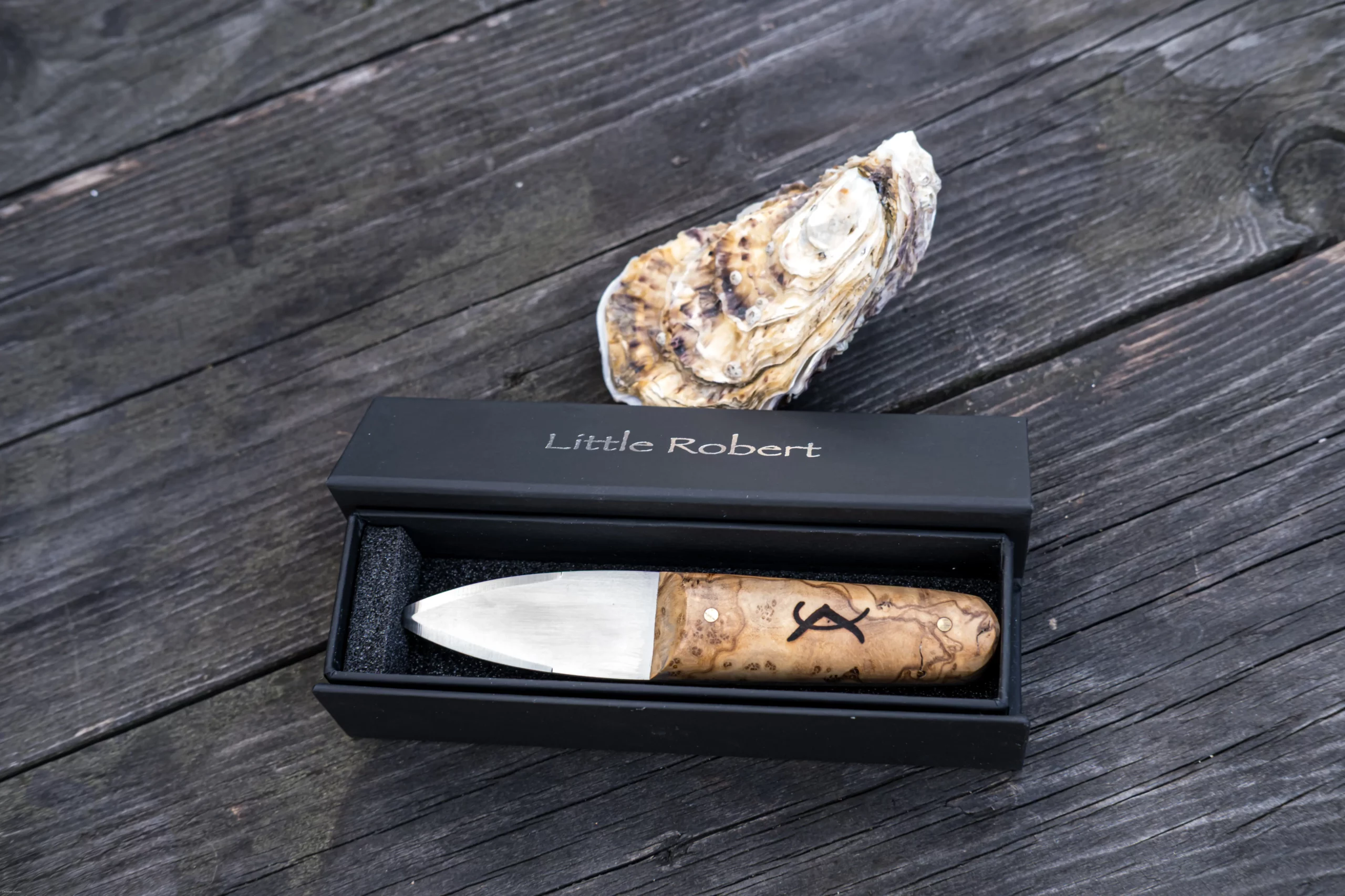 Couteau à huîtres le Little Robert dans son coffret noir, posé sur une table en bois à côté d'une huître.